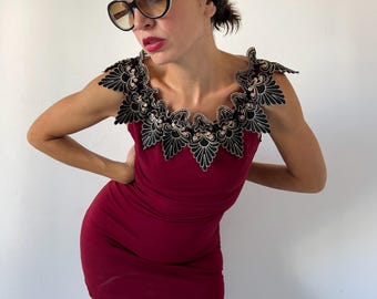 Puede incluir: Un vestido corto sin mangas de color burdeos con un escote ancho y festoneado, adornado con intrincados bordados en negro y dorado. El detalle decorativo del cuello se extiende sobre los hombros, creando un contraste con la tela roja oscura.