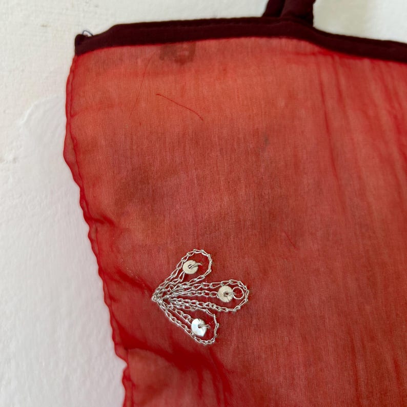 May include: A sheer red fabric bag featuring a dark red trim along the top edge. The bag is decorated with a small, silver-toned metallic embellishment shaped like a fan or leaf, which is accented with three small, circular silver sequins.