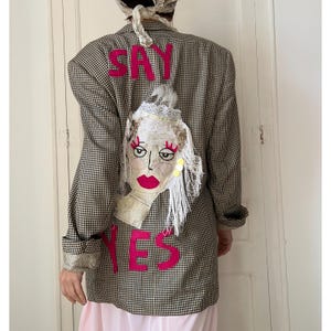 May include: A brown and black gingham blazer featuring the hot pink text SAY YES on the back. An embroidered face appliqué with white lace, fringe, and sequins is centred between the words. The blazer has long sleeves and a structured fit.