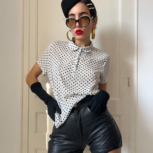 Puede incluir: Una camisa blanca con lunares negros y cuello se combina con un pantalón corto de cuero negro. El conjunto se completa con una boina negra, gafas de sol, pendientes de aro dorados y guantes negros. El estilo general es de inspiración vintage.