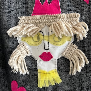 May include: A dark gray plaid fabric featuring a textile applique of a face. The face has a pink felt crown, cream-colored yarn hair, yellow mesh glasses, embroidered black facial features, pink lips, and a yellow mesh collar detail.