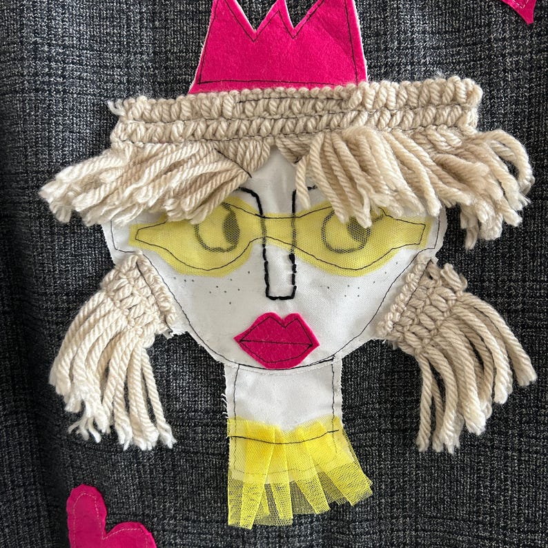 May include: A dark gray plaid fabric featuring a textile applique of a face. The face has a pink felt crown, cream-colored yarn hair, yellow mesh glasses, embroidered black facial features, pink lips, and a yellow mesh collar detail.