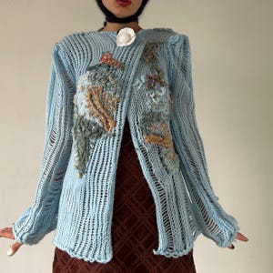 May include: A light blue open-knit cardigan featuring long sleeves with vertical ladder-stitch detailing, an asymmetrical front closure, and textured, multi-colored yarn embroidery accents. A single white floral-shaped button secures the neckline.