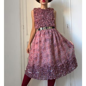 May include: A sleeveless midi dress in a dusty rose and mauve floral print, featuring a bodice and hemline covered in dense, ruffled fabric layers. The dress is cinched at the waist with a black belt displaying the gold-toned text MOSCHINO.