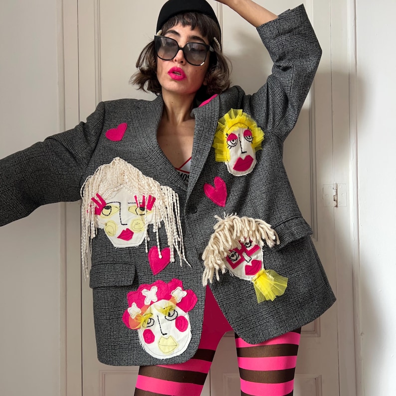 May include: A charcoal gray plaid blazer featuring multiple embroidered fabric face appliqu&eacute;s with yarn hair and pink heart patches. The blazer has a notched lapel, two front flap pockets, and a single button closure, worn over pink and brown striped leggings.