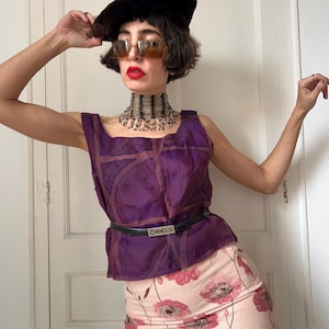 Könnte beinhalten: Ein ärmelloses violettes Oberteil mit dunklem geometrischem Gittermuster, das mit einem dünnen schwarzen Gürtel mit silberner Moschino-Schnalle in der Taille gerafft ist. Das Outfit umfasst einen hellrosa Rock mit dunkelrotem Blumenmuster, ein braunes Barett und eine Sonnenbrille.