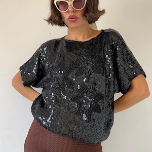 Può includere: Top nero con paillettes, maniche corte e scollatura rotonda. La parte superiore presenta tre grandi disegni floreali, anch'essi in paillettes nere. L'indumento è indossato con pantaloni marroni e occhiali da sole.