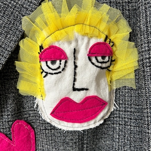 May include: A gray tweed blazer featuring a large, hand-stitched fabric applique of a face. The face has bright yellow tulle hair, hot pink felt eyelids and lips, and black embroidered outlines for the eyes, nose, and facial structure.