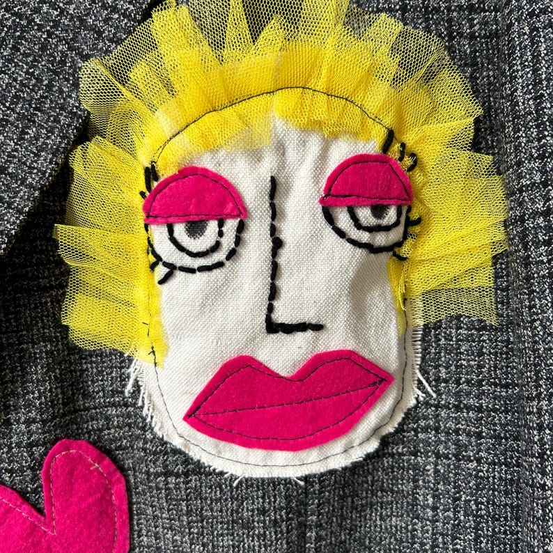 May include: A gray tweed blazer featuring a large, hand-stitched fabric applique of a face. The face has bright yellow tulle hair, hot pink felt eyelids and lips, and black embroidered outlines for the eyes, nose, and facial structure.