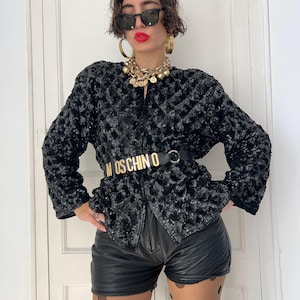 May include: A black sequined jacket with a textured pattern, paired with black leather shorts and a gold belt with the word "MOSCHINO" on it. The jacket is zipped up. The model is wearing sunglasses and gold jewellery.