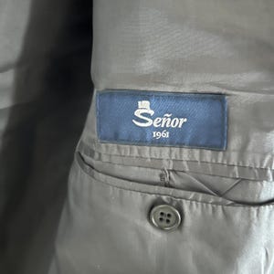 May include: A dark gray interior lining of a garment featuring a rectangular blue fabric label stitched above an interior pocket. The label displays the text Se&ntilde;or 1961 in white lettering with a small top hat icon above the word Se&ntilde;or.