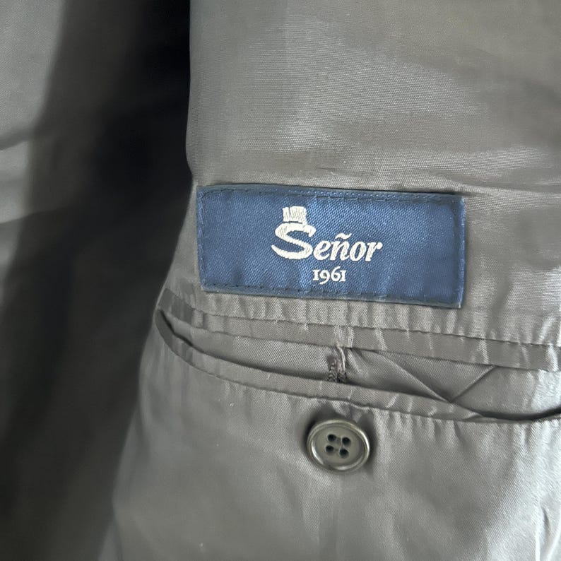 May include: A dark gray interior lining of a garment featuring a rectangular blue fabric label stitched above an interior pocket. The label displays the text Se&ntilde;or 1961 in white lettering with a small top hat icon above the word Se&ntilde;or.