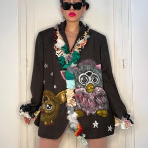 Puede incluir: Blazer marrón oscuro con detalles de estrellas y dos apliques de personajes Furby. El blazer presenta un cuello y puños con volantes de colores. Los personajes Furby son en tonos rosa, gris y amarillo.