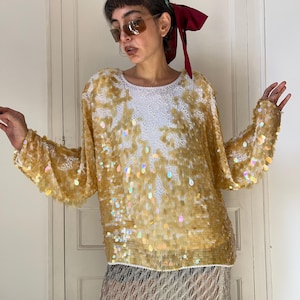 Puede incluir: Una blusa de manga larga con base blanca, intrincado bordado blanco y una densa capa de lentejuelas brillantes en forma de lágrima de color dorado, iridiscente y translúcido que crean una superficie texturizada y reflectante.
