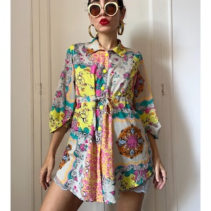 May include: A button-down shirt featuring a vibrant, multi-colored pattern with shades of yellow, pink, teal, and white. The garment includes a collar, three-quarter length sleeves, and a drawstring waist tie, showcasing a complex, ornate design.