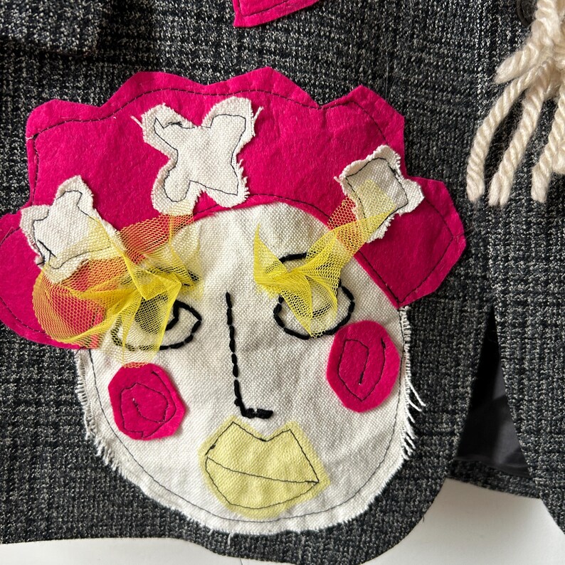 May include: A dark gray plaid blazer featuring a large, hand-stitched fabric applique of a face. The face has a bright pink hat, yellow tulle eyelashes, pink circular cheeks, and a pale yellow mouth, all attached to the textured wool-like fabric.