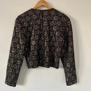 May include: A long-sleeved jacket featuring a dark brown base color with an intricate, repeating metallic silver floral pattern throughout. The garment has a round neckline, a straight hemline, and is displayed on a wooden hanger.