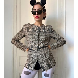 May include: A gray and white paisley-patterned jacket with a black belt. The jacket has a button-up front and long sleeves. The belt has a silver buckle with the word "MOSCHINO" on it. The jacket is paired with black shorts and white tights with black polka dots.