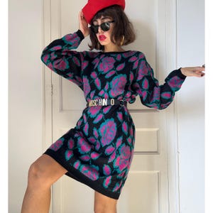 Peut inclure: Une robe-pull noire ornée d'un motif répétitif de roses magenta et bleu sarcelle éclatantes. La robe présente des manches longues, une encolure ronde et est cintrée à la taille par une ceinture en cuir noir avec l'inscription MOSCHINO en métal doré.