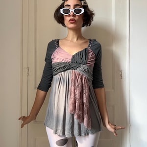 May include: A three-quarter sleeve top featuring a charcoal gray bodice with a twisted front detail, a dusty pink fabric overlay, and a vertical panel of matching pink lace. The garment includes dark gray sleeves and a flared, light gray hem.