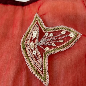May include: A sheer red fabric featuring an embroidered leaf-shaped applique. The design includes concentric borders of light pink and olive green thread, accented with silver metallic stitching and small, circular silver sequins.