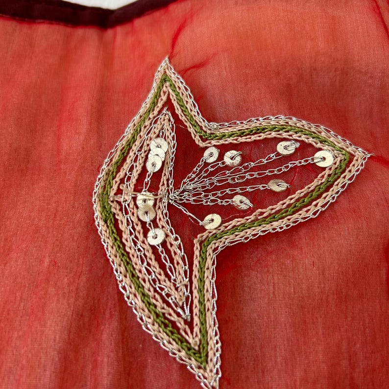 May include: A sheer red fabric featuring an embroidered leaf-shaped applique. The design includes concentric borders of light pink and olive green thread, accented with silver metallic stitching and small, circular silver sequins.