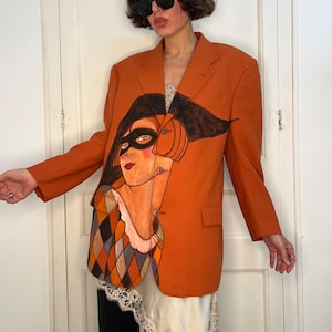 Peut inclure: Un blazer orange orné d'une illustration peinte à la main représentant un personnage portant un masque noir et un vêtement à motif arlequin. Le blazer possède des manches longues, un revers cranté, trois boutons et deux poches à rabat.