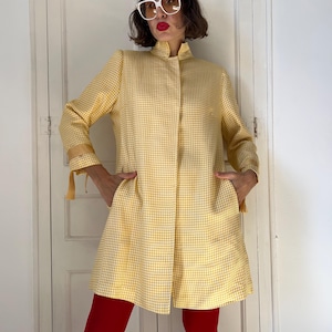 May include: A yellow and white houndstooth pattern coat with a mandarin collar and long sleeves. The coat has two pockets and a slight A-line shape. The sleeves have a decorative tie detail at the cuffs. The person is wearing red pants.