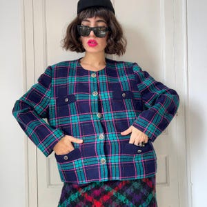 Pode incluir: Uma jaqueta xadrez azul marinho, verde-azulado e rosa com botões prateados e bolsos. A jaqueta faz parte de um conjunto a condizer com uma saia xadrez. A jaqueta tem decote redondo e é abotoada. A roupa é vintage.