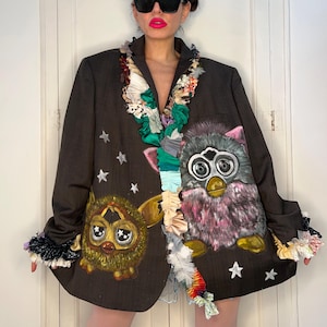 May include: A dark brown blazer featuring hand-painted Furby characters and white star accents. The blazer has decorative fabric trim around the collar and cuffs, adding a unique, artistic flair to the garment. The design is whimsical and eye-catching.