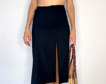 Pode incluir: Uma saia midi preta sólida com design de cintura alta e uma fenda vertical proeminente no centro da frente que se estende da bainha até a metade da coxa, confeccionada em tecido liso e opaco com uma silhueta reta e limpa.