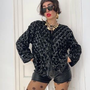 May include: A black, textured jacket with a quilted pattern. The jacket has long sleeves and a zipper closure. The model is wearing black shorts, sunglasses, and gold jewelry. The background is a white door.