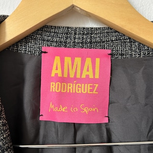 May include: A charcoal gray blazer with a textured, fine-check pattern, featuring a bright pink interior label with yellow text that reads AMAI RODR&Iacute;GUEZ and Made in Spain, hanging on a wooden hanger against a plain white background.