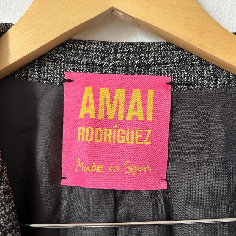 May include: A charcoal gray blazer with a textured, fine-check pattern, featuring a bright pink interior label with yellow text that reads AMAI RODR&Iacute;GUEZ and Made in Spain, hanging on a wooden hanger against a plain white background.