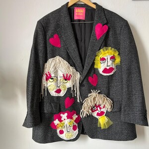May include: A dark gray plaid blazer featuring a bright pink label that reads AMAI RODRIGUEZ. The garment is decorated with four distinct fabric face appliqu&eacute;s with yarn hair and tulle accents, interspersed with several bright pink fabric hearts.