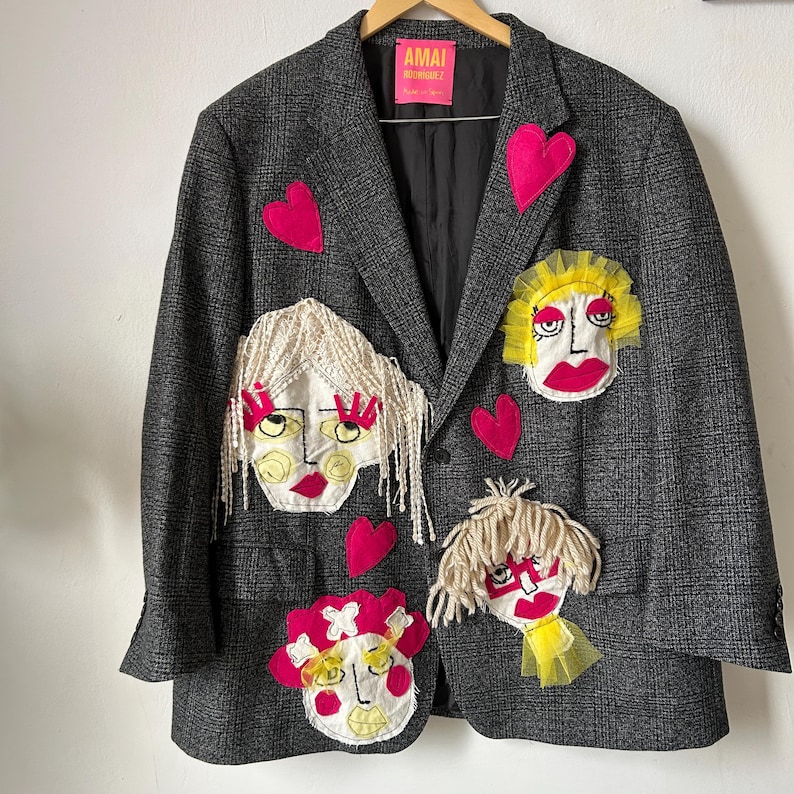 May include: A dark gray plaid blazer featuring a bright pink label that reads AMAI RODRIGUEZ. The garment is decorated with four distinct fabric face appliqu&eacute;s with yarn hair and tulle accents, interspersed with several bright pink fabric hearts.