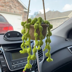 May include: A crocheted hanging plant with green leaves and a brown pot. The plant is hanging from a rearview mirror in a car.