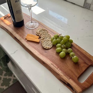 May include: A wooden serving board with a natural edge, holding a bottle of wine, a wine glass, crackers, cheese, and green grapes. The board has the text "uncork & unwind" engraved on it. The board has a handle on each end.