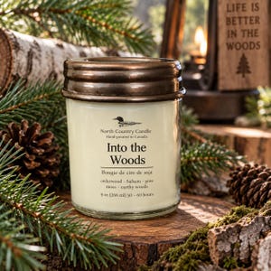 May include: A clear glass candle jar with a metallic bronze lid. The label features a loon icon, the text North Country Candle, Into the Woods, Bougie de cire de soja, cedarwood, balsam, pine, moss, earthy woods, and 9 oz (266 ml) 50-60 hours.