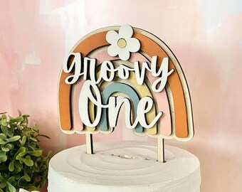 May include: A white cake with a rainbow and flower topper that says "groovy one".