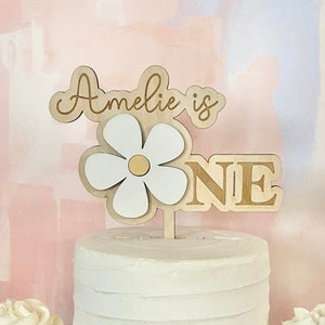 Pu&ograve; includere: Una torta bianca glassata con un cake topper a forma di margherita in legno che recita "Amelie is ONE". Due cupcakes con glassa bianca si trovano su entrambi i lati della torta. Lo sfondo &egrave; un design astratto rosa e blu pastello.