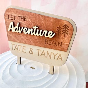 May include: Wooden cake topper with the words "Let the Adventure Begin" and the names "Tate & Tanya". The topper has a brown top section and a light-colored bottom section, with two small posts for insertion into the cake.