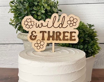 May include: Wooden cake topper with the text "wild & three" and small flower designs. The topper is on a white frosted cake.