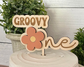 May include: A wooden cake topper with the words "Groovy One" in a retro font. The topper features a pink flower with a yellow center and a cursive "one" in brown.