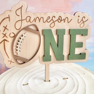 May include: A wooden cake topper with the text "Jameson is ONE." The topper features a football and play diagram design. The word "ONE" is in green, and the other elements are in natural wood tones. The topper is inserted into a white frosted cake.