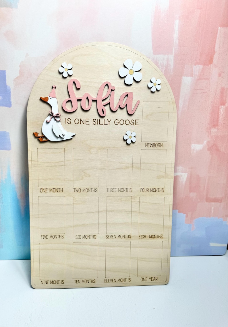 May include: Wooden milestone board with the name "Sofia" in pink script, a cartoon goose, and flower accents. The board is designed to track a baby's first year, with spaces for each month and "Newborn" at the top.