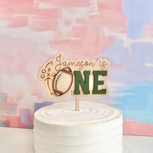 May include: A wooden cake topper with the text "Jameson is ONE" and a football design. The word "ONE" is in green, and the topper is placed on top of a white frosted cake. The background has pink and blue brushstrokes.