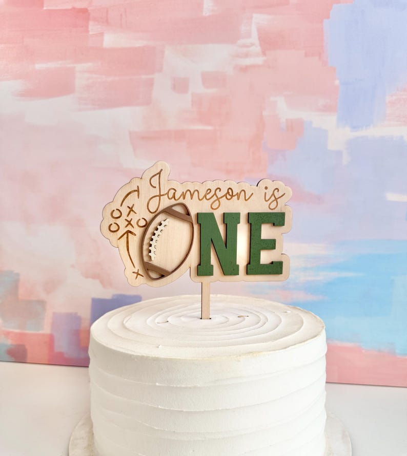 May include: A wooden cake topper with the text "Jameson is ONE" and a football design. The word "ONE" is in green, and the topper is placed on top of a white frosted cake. The background has pink and blue brushstrokes.