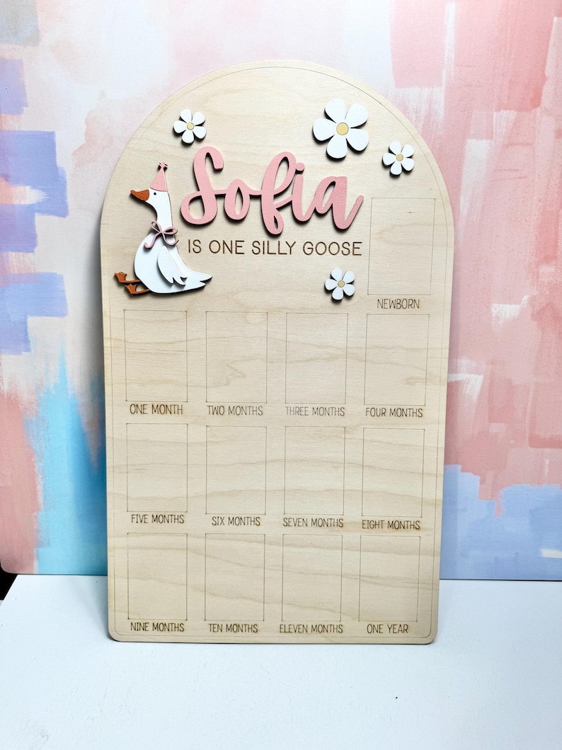 May include: Wooden milestone board with the name "Sofia" in pink script, a cartoon goose, and flower accents. The board is labeled with months from newborn to one year, perfect for tracking a baby's growth.