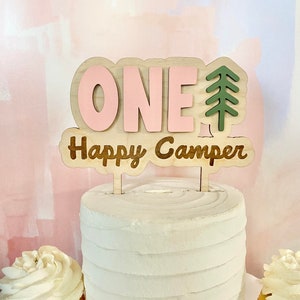 K&ouml;nnte beinhalten: Ein Holz-Kuchentopper mit dem Wort "ONE" in rosa Buchstaben und den Worten "Happy Camper" in braunen Buchstaben. Rechts neben dem Wort "ONE" befindet sich eine gr&uuml;ne Baumsilhouette.